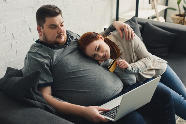 Korkea kulma näkymä ylipainoinen poikaystävä ja tyttöystävä ostoksia verkossa kannettavan tietokoneen ja luottokortin kotona
 - Valokuva, kuva