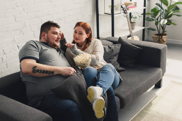 size plus girlfriend feeding boyfriend with popcorn on sofa at home - Фото, изображение