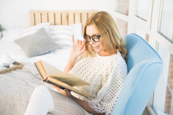 Young blond woman at home sitting on modern chair in front of window relaxing in her living room reading book  - Foto, Imagem