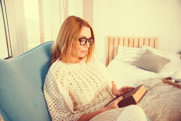 junge blonde Frau sitzt zu Hause auf modernem Stuhl vor Fenster und entspannt sich in ihrem Wohnzimmer beim Lesen von Büchern  - Foto, Bild