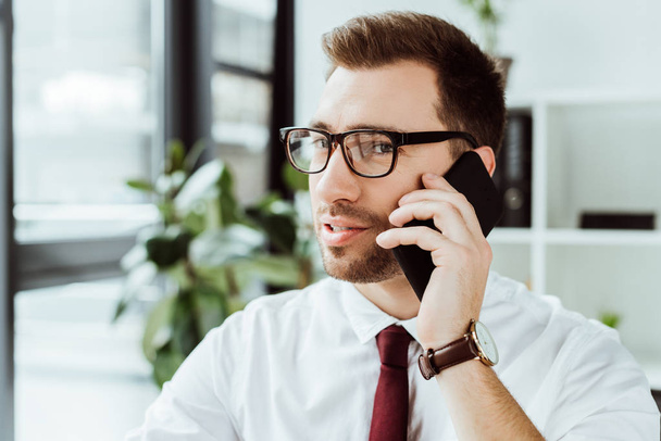 красивый бизнесмен в очках разговаривает на смартфоне в офисе
 - Фото, изображение