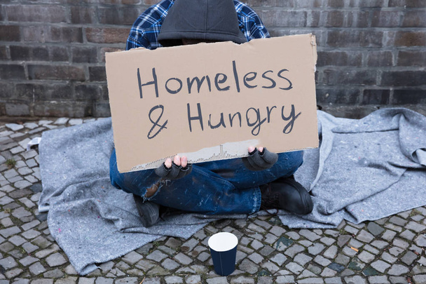 Male Beggar Lying On Street With Homeless And Hungry Text On Cardboard - Foto, Imagen