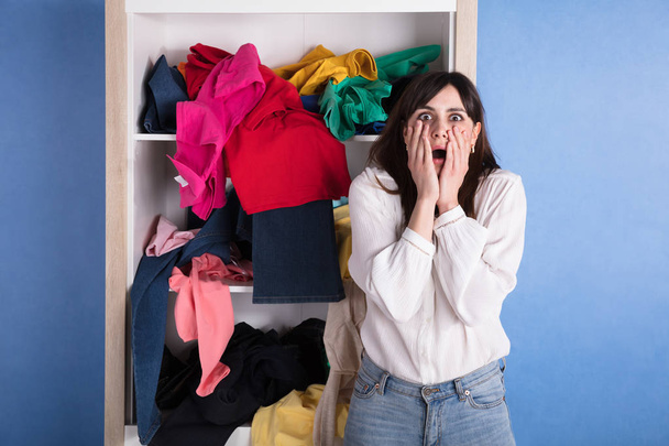 Portrait Of A Shocked Woman In Front Of Scattered Clothes On Shelf - 写真・画像
