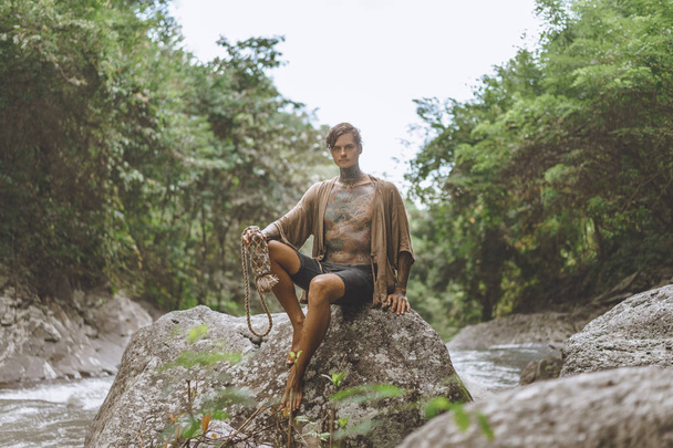 uomo tatuato con borraccia poggiata su roccia con piante verdi e fiume sullo sfondo, Bali, Indonesia
 - Foto, immagini