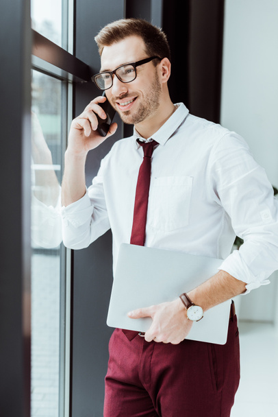 businessman talking on smartphone and holding laptop in modern office - Φωτογραφία, εικόνα