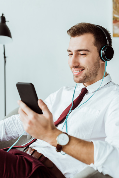 χαμογελώντας επιχειρηματίας ακούγοντας μουσική στα ακουστικά και χρησιμοποιώντας το smartphone - Φωτογραφία, εικόνα