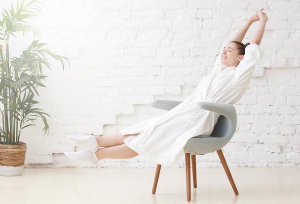 Horizontal shot of young smiling woman in white bathrobe stretching in the morning after waking up - 写真・画像