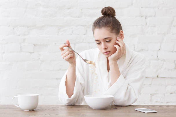 Young woman with no appetite, tired, bad mood and sleepy, sitting in the white loft kitchen, having unsavory unpalatable unappetizing breakfast - Photo, Image