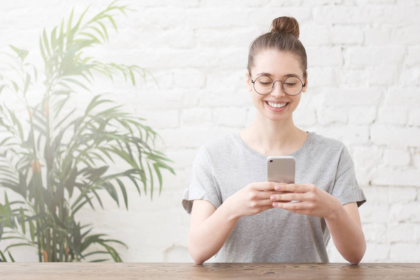 Smiling young female entrepreneur in grey t-shirt working online with smartphone while sitting at wooden table in her home office - Foto, Imagen