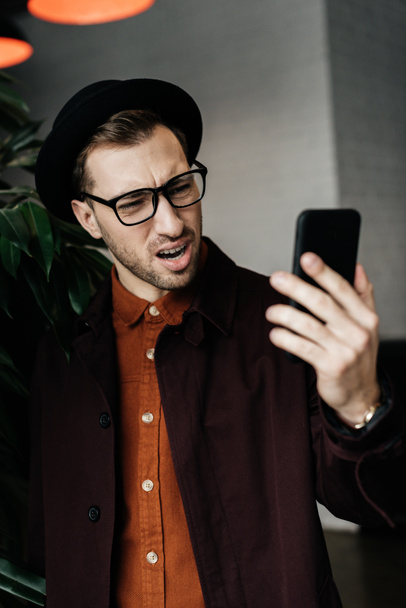 handsome stressed man in hat and glasses looking at smartphone - Photo, Image