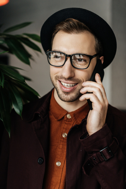 handsome cheerful man in eyeglasses and hat talking on smartphone - Fotó, kép