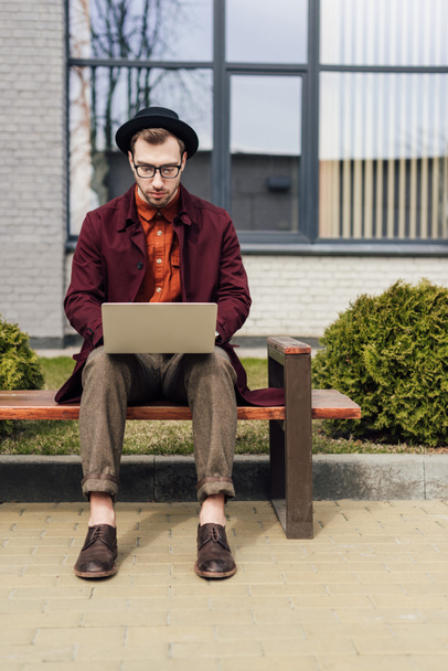 handsome fashionable man using laptop and sitting on bench - Фото, зображення