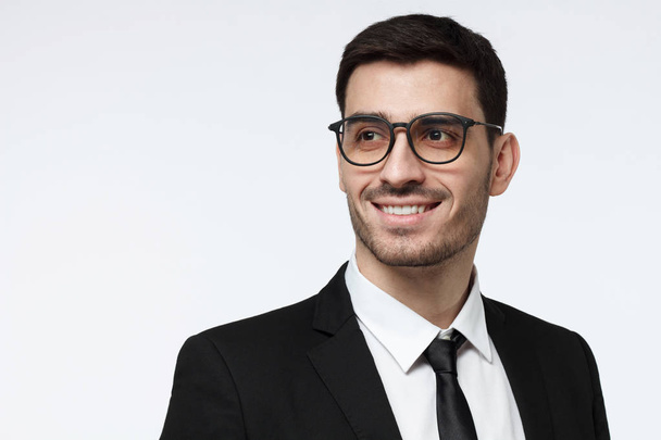 Closeup shot of young corporate advisor in trendy spectacles pictured isolated on grey background wearing formal black suit, showing toothy open smile, looking to the left, positive and confident - Foto, Imagem