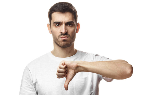 Disgruntled grumpy dissatisfied annoyed unhappy young man showing thumb down as dislike gesture isolated on white background - Photo, image