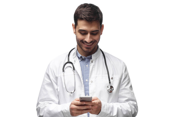 Closeup photo of man doctor standing isolated on white background looking attentively at screen of cellphone, smiling nicely - Foto, afbeelding