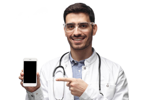 Studio closeup of positive man doctor isolated on white background standing in uniform with stethoscope around neck showing blank cellphone screen with copyspace, smiling happily - Photo, Image