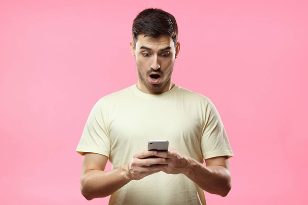 Close up portrait of handsome young man in beige t-shirt, holding smartphone, looking scared and confused reading text message or e-mail - Φωτογραφία, εικόνα