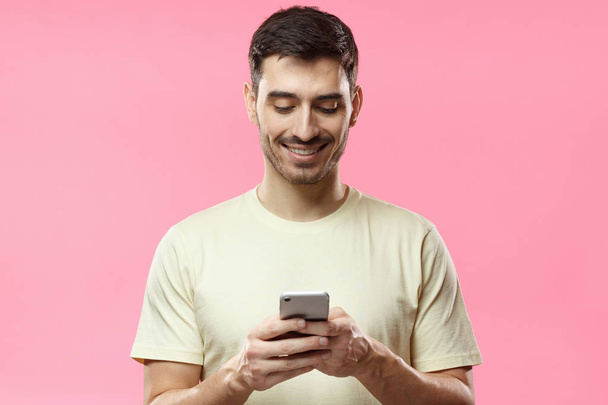 Closeup photo of man in beige tshirt standing isolated on pink background looking attentively at screen of cellphone, browsing web pages and smiling nicely while chatting with friends - Photo, image