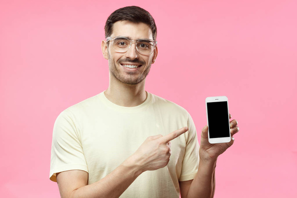 Handsome man in beige tshirt in transparent glasses isolated on pink background, presenting smart phone and pointing with finger at blank black screen with copyspace for ads - Foto, Imagem