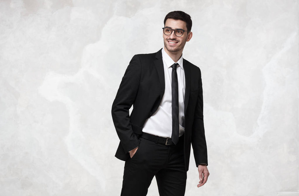 Elegant smiling young business man walking against grey texture wall - Foto, afbeelding