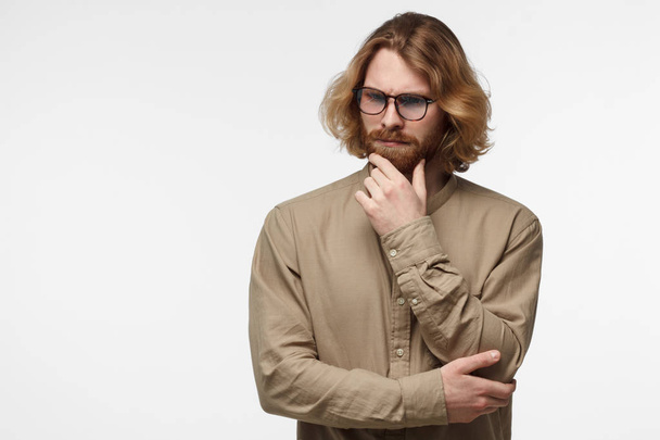 Thoughtful young bearded man in olive shirt, holding his chin with hand, solving relationship problem. Doubt concept.  - 写真・画像
