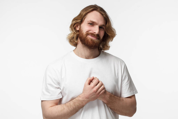 Kind hearted friendly nice bearded guy shows kindness, wearing blank white tshirt. Portrait of smiling positive young man keeps hands on chest, expresses sympathy  - Foto, Imagen