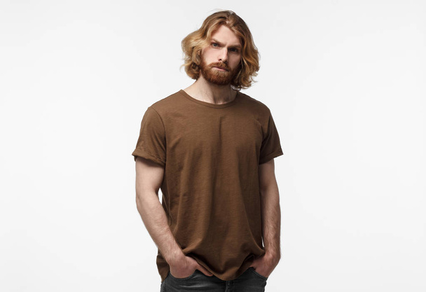 Young brutal bearded man with long hair standing with hands in pockets, in brown tshirt, isolated on grey background - Fotó, kép