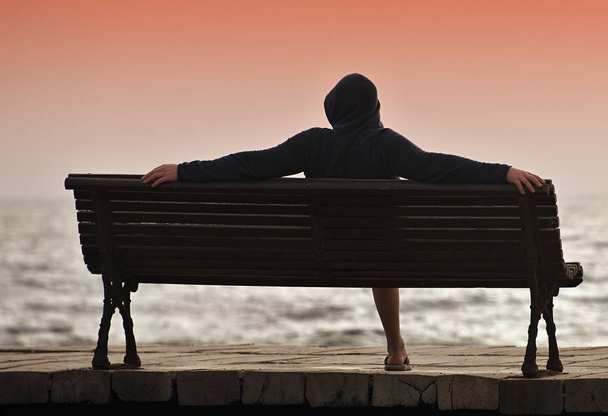 Een jonge man met een kap die zittend op een bankje met uitzicht op de Oceaan - Foto, afbeelding