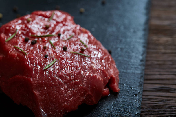 Raw meat beef steaks on black slate board with spices and rosemary over wooden background, copy space - Фото, зображення