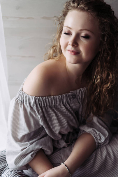 young attractive curly girl sitting by the window, enjoying rest and smiling, eyes closed - Fotoğraf, Görsel