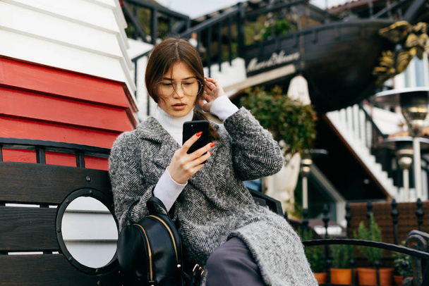 stylish dark-haired girl in a trendy gray coat sits on a bench outdoors and looks in her smartphone - Photo, Image