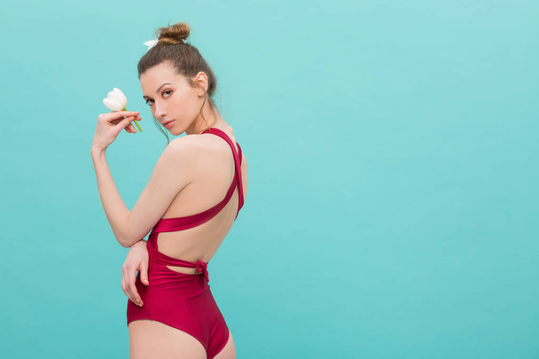 Slim sexy model in a red wine color body swimsuit holding white tulip flower and posing over mint colour background. Skinny girl in swimwear looking fresh and hot - Фото, изображение