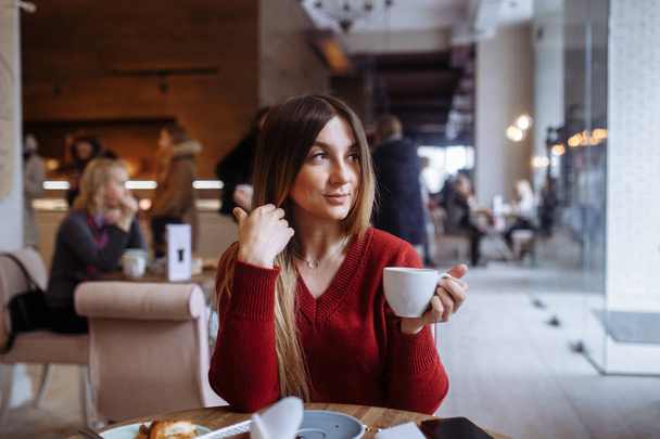 Beautiful young woman holding coffee cup sitting at table in cafe - Фото, изображение