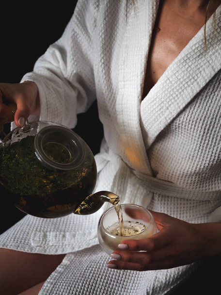 Girl in a white bath robe puiring herbal tea in a transparent glass - Zdjęcie, obraz