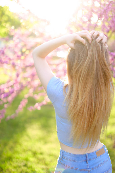 Blonde vrouw permanent in de stralen van de zon met groen gras en bloesem achtergrond. - Foto, afbeelding