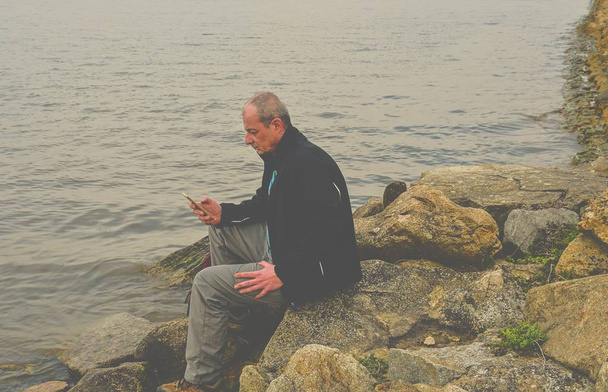 Midle aged man sitting on the shore of the lake. Solitude mature man using mobile phone on the bank. Concept of activ man - Zdjęcie, obraz