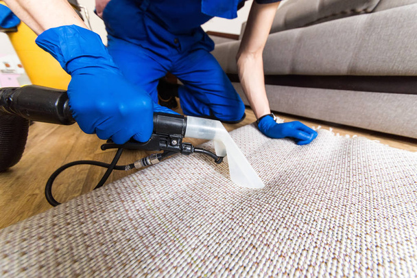 Cleaning service. Man janitor in gloves and uniform vacuum clean white carpet with professional equipment - Foto, afbeelding