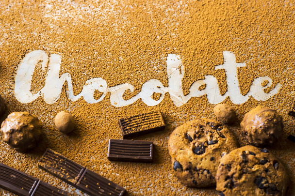 The word chocolate printed on cocoa on a gray background among cocoa and chocolate chip cookies, chocolate pieces, nuts in chocolate and sweets. I love chocolate concept - Photo, Image