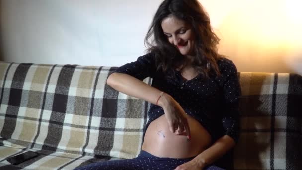 Ευτυχισμένος έγκυος γυναίκα στηρίζεται σε έναν καναπέ και χαϊδεύοντας την κοιλιά της. - Πλάνα, βίντεο