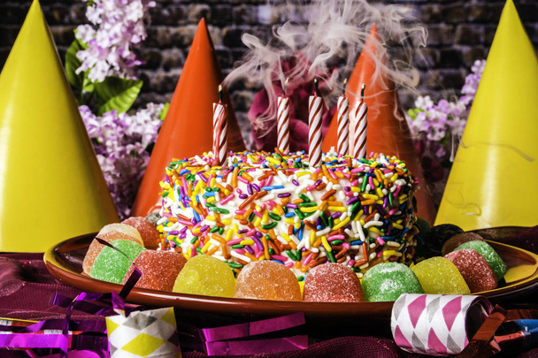 birthday cake with rainbow sprinkles on red plate surrounded with assorted gumdrops with smoking candles party hats party favors and spring flowers with brick wall background - Photo, Image