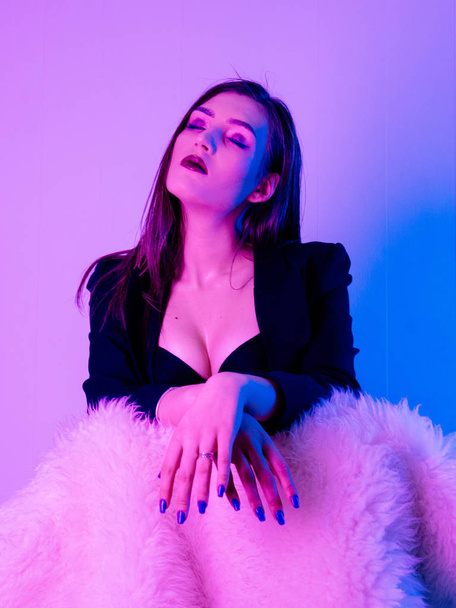 Sexy Beautiful Woman wearing jacket sitting on fur chair in ultraviolet light - Photo, Image
