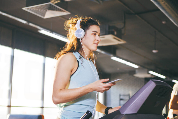 Theme sport and music. A beautiful Caucasian woman running in gym on treadmill. On head big white headphones, the girl listens to music during a cardio workout for weight loss and uses phone in hand - Photo, Image