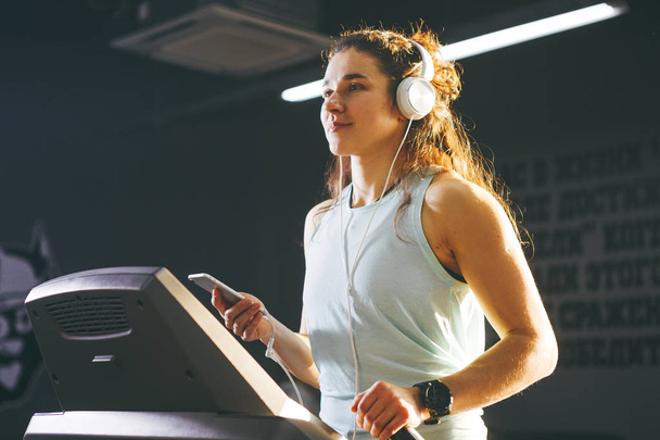Thema Sport und Musik. eine schöne kaukasische Frau, die im Fitnessstudio auf dem Laufband läuft. auf dem Kopf große weiße Kopfhörer, das Mädchen hört Musik während eines Cardio-Workouts zur Gewichtsabnahme und benutzt das Handy in der Hand - Foto, Bild