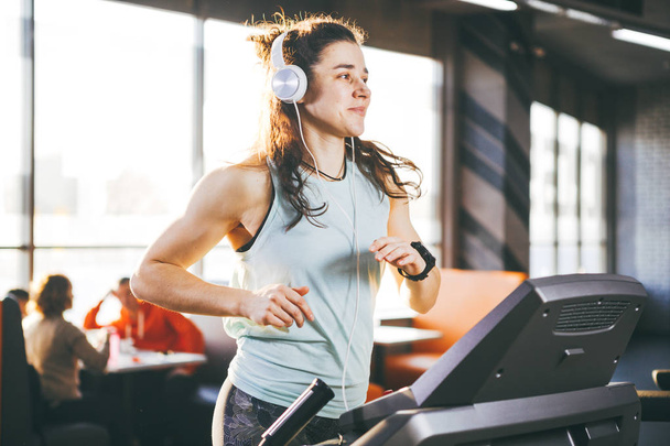 Theme is sport and music. A beautiful inflated woman runs in the gym on a treadmill. On her head are big white headphones, the girl listens to music during a cardio workout for weight loss - Foto, imagen