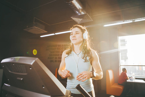 Το θέμα είναι άθλημα και μουσική. Μια όμορφη γυναίκα φουσκωμένα τρέχει στο γυμναστήριο σε ένα διάδρομο. Στο κεφάλι της είναι μεγάλο λευκά ακουστικά, το κορίτσι που ακούει τη μουσική κατά τη διάρκεια της προπόνηση cardio για απώλεια βάρους - Φωτογραφία, εικόνα