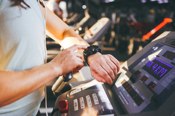 Close-up of a young Caucasian woman's hand in the gym uses a sports watch, a black pulse wrist on the back of a treadmill in a spotlight in sunny weather - Foto, Imagen