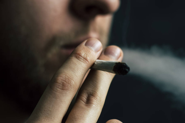 raucht Cannabiskraut, einen Joint und ein Feuerzeug in seinen Händen. Rauch auf schwarzem Hintergrund. Konzepte des medizinischen Marihuana-Konsums und der Legalisierung von Cannabis. - Foto, Bild