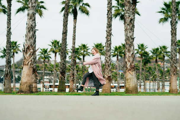 happy woman in a stylish pink coat walking in a park next to green palms, enjoying spring and warmth - Photo, Image