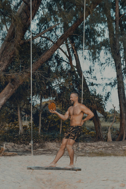 Молодой красивый мужчина стоит на деревянных качелях на пляже с панданскими фруктами в руке. Без рубашки в шортах, мускулистых телах. Остров Ронг Самлум, пляж Лэзи. Камбоджа, Азия
 - Фото, изображение