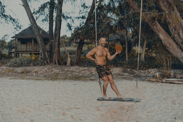 Молодой красивый мужчина стоит на деревянных качелях на пляже с панданскими фруктами в руке. Без рубашки в шортах, мускулистых телах. Остров Ронг Самлум, пляж Лэзи. Камбоджа, Азия
 - Фото, изображение